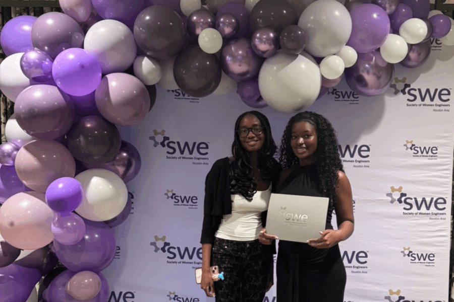 Two young Black women, dressed in black clothing, stand in front of a light purple backdrop, with a cascade of balloons in shades of purple framing them on the top and left. Temi Ilori, standing on the right, holds a certificate at waist height.