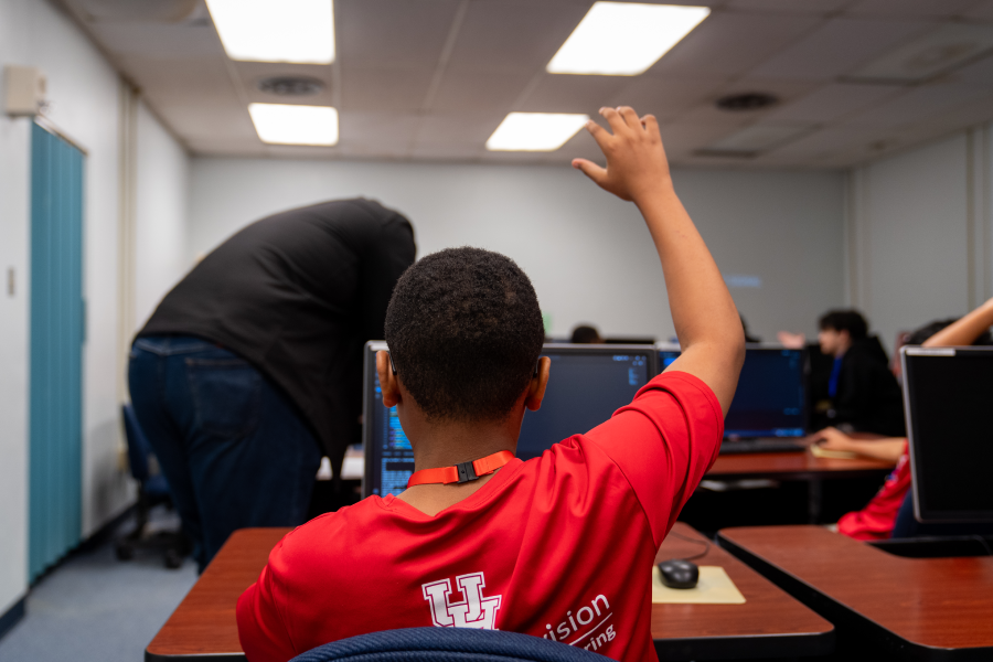 A middle-grad child sits at a computer table in a room with other campers, raising his right hand in the air.