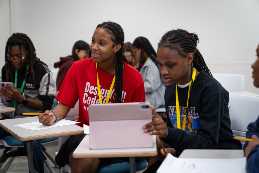 Two middle-grade students sit at desks in a room with other students. One is wearing a red t-shirt, and the other is looking at the screen of a light pink tablet on the desk in front of her. 