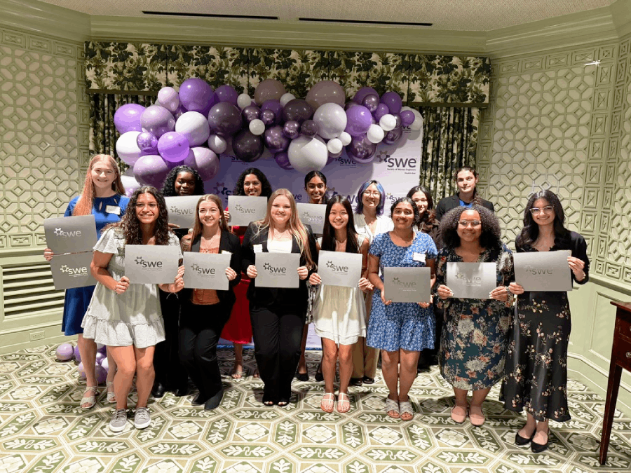 Two rows of of college-aged women stand in front of a light purple backdrop with a cascade of balloons in shades of purple across the top of the backdrop. They are each holding a certificate. Ilori stands second from left in the backmost row.