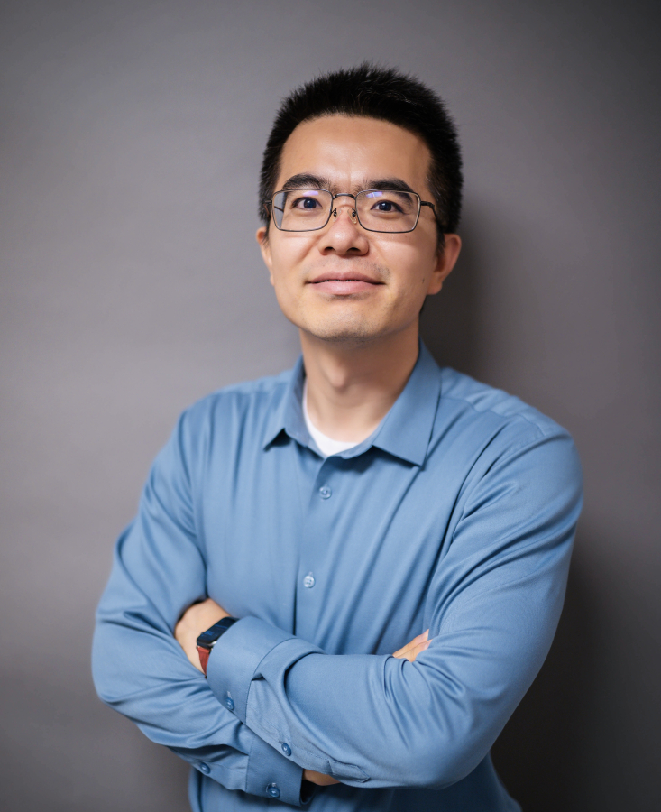 Bo Zhao, smiling, wears a blue collared shirt and is standing in front of a grey background with his arms crossed.