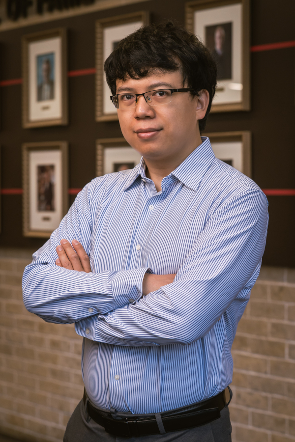 Lihong Zhao, wearing a blue long-sleeved dress shirt, stands in front of a row of wall portraits. His arms are crossed in front of him.