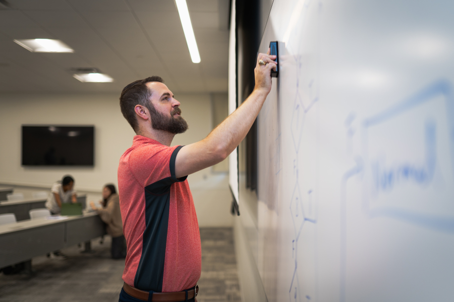Zelisko reaches up to erase part of a dry-erase board in a lecture hall. There is blue writing on the board.