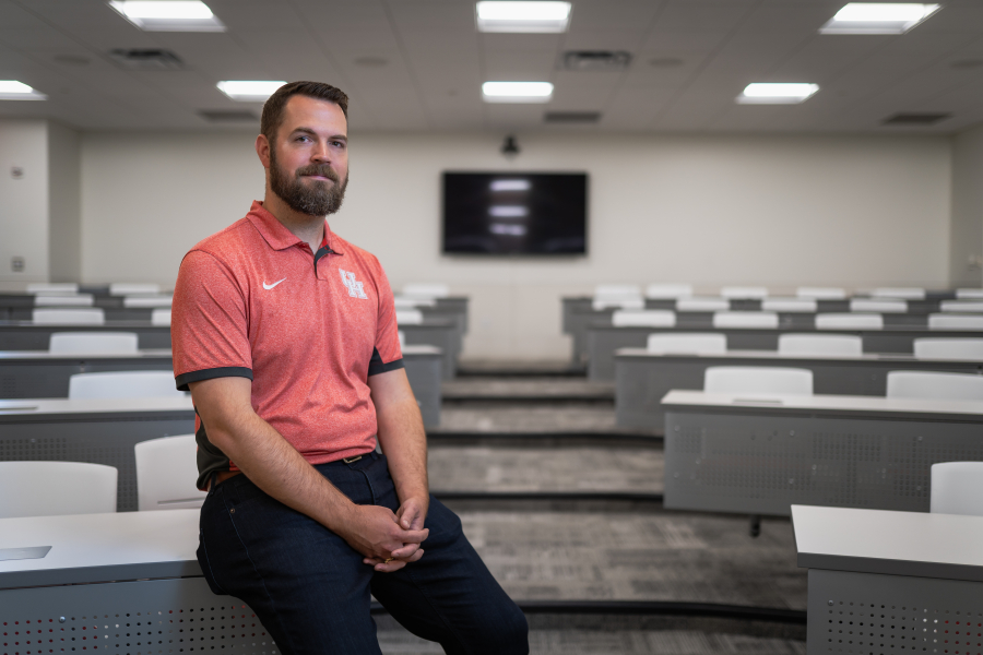 Matt Zelisko, wearing a red UH polo shirt, sits at the edge of a grey lecture hall table.