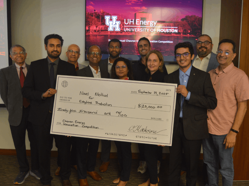 Imam and Pandit stand on either side of an oversized white check, holding it between them. They are surrounded by stakeholders and competition evaluators.