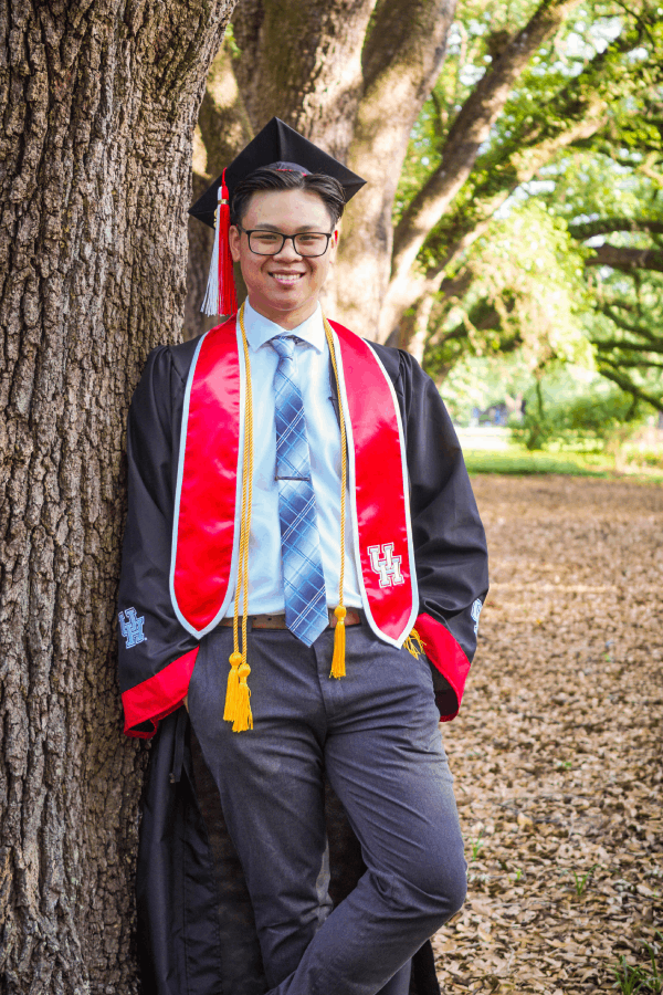 Nathan Tran, a young Asian man dressed in graduation regalia, smiles and stands against a tree. He is wearing a black mortarboard cap with two tassels, a red and white stole, and a black gown over a charcoal suit and striped tie.