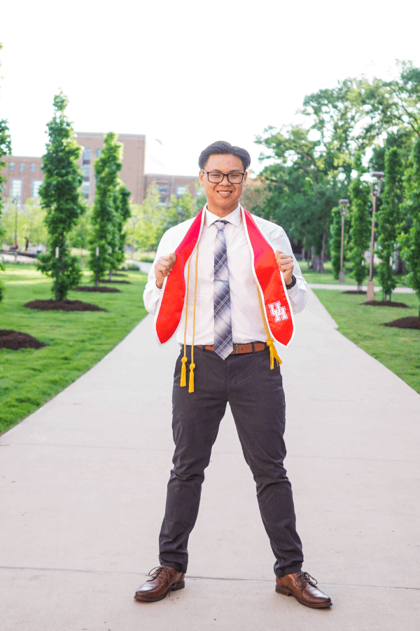 Nathan Tran stands upright on an outdoor path flanked by trees, wearing a red graduation stole over a light-colored dress shirt and striped tie.