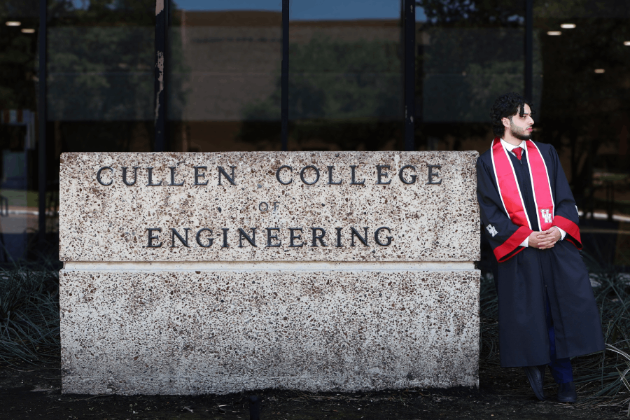 Abdel-Khalek, wearing graduation regalia, stands beside the carved stone sign reading "Cullen College of Engineering" outside of the college's building on the main campus.
