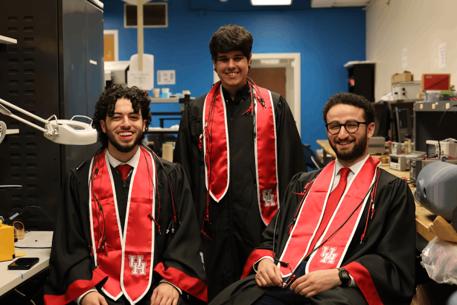 Three people in graduation regalia, including red and white stoles, sit together in the middle of a lab on campus. The wall behind them is deep blue. Abdel-Khalek sits on the far left of the photo.