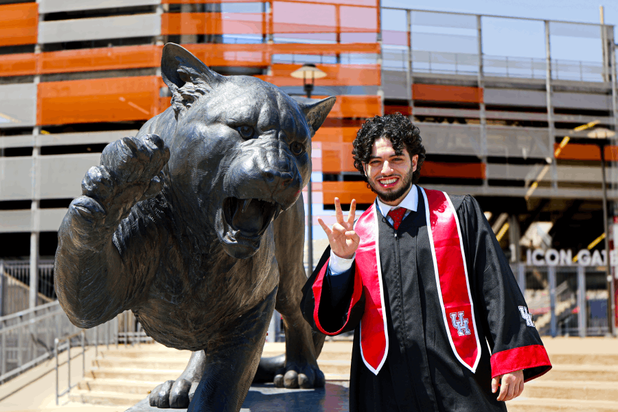 Abdullah H. Abdel-Khalek, dressed in graduation regalia, stands to the right of the bronze Shasta I statute outside of TDECU Stadium while holding up the UH cougar paw handsign.