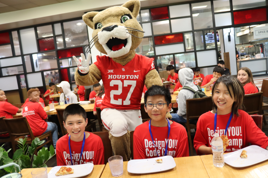 Three middle-grade children in red t-shirts sit at a lunch table. Behind them stands UH mascot Shasta.