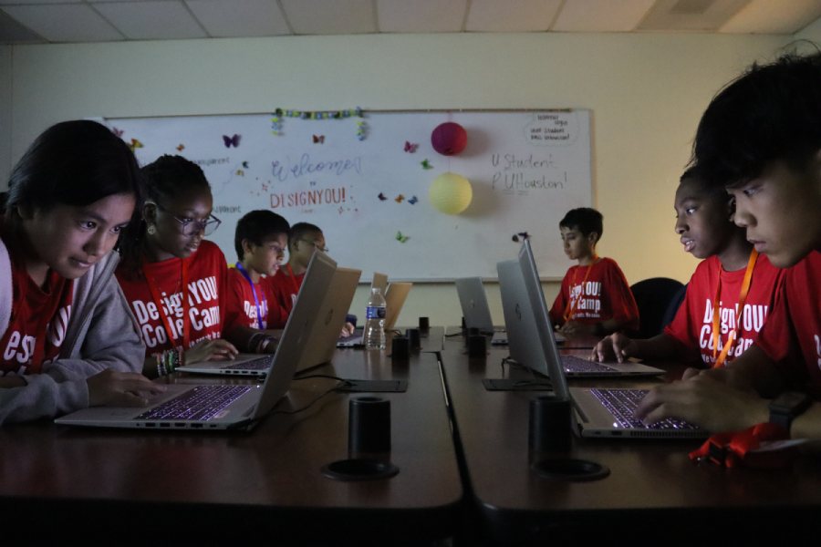Several middle-grade campers sit on either side of a table in a darkened room. They each work on a laptop sitting in front of them.