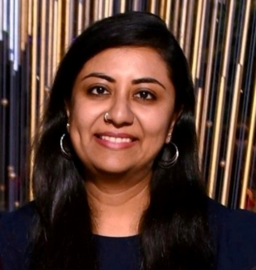 Nirathi Keerthi Govindu, a woman with long, dark hair and olive skin, smiles at the camera in front of a gold and black background. 