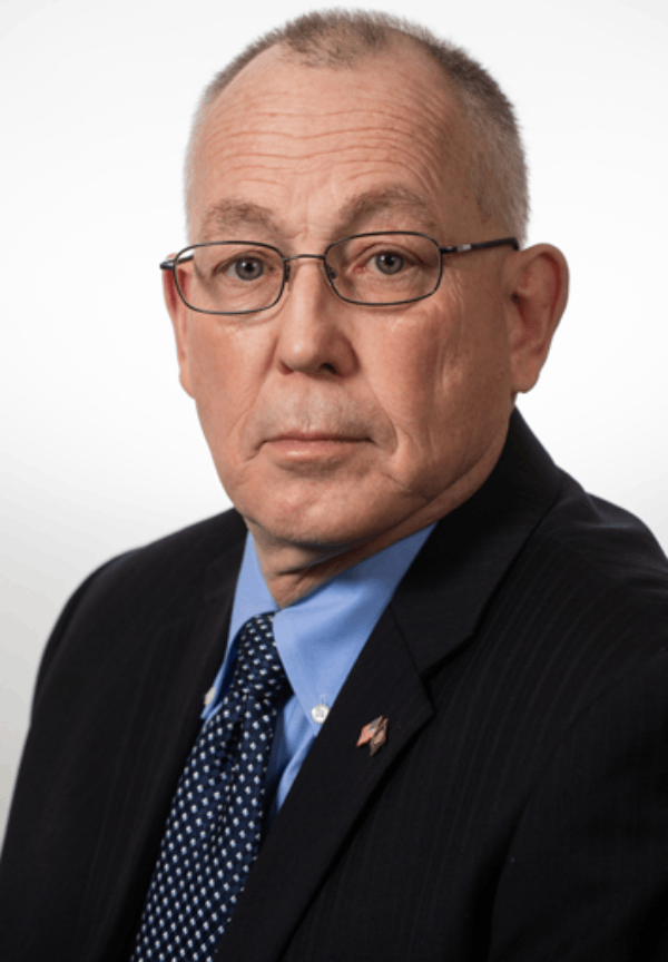 A headshot of Lawrence Schulze, who looks directly at the camera, in a black suit jacket and a blue dress shirt and tie. Schulze also wears glasses and has a short, grey haircut.