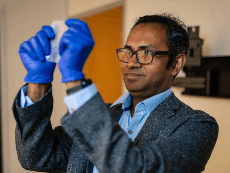 Rahmnan, wearing a glasses and a light blue shirt under a grey suit jacket, holds a piece of translucent bioplastic up to eye level. He is wearing blue gloves.