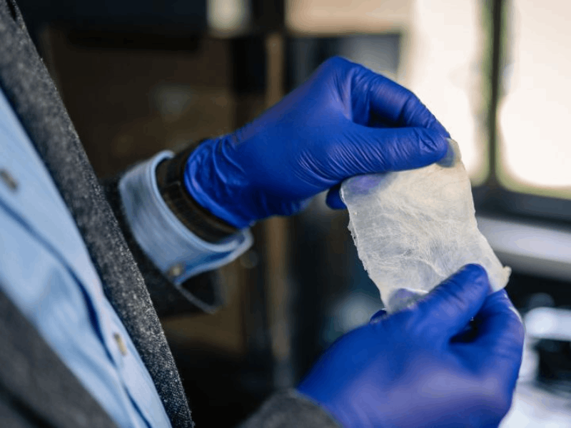 Two gloved hands, blue, hold a small, rectangular piece of translucent white bioplastic between them,