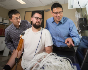 With high-density electrodes placed on the arm of grad student Nick Dias, Yingchun Zhang (far right) monitors muscle contractions to pinpoint the neuromuscular junction. Grad student Chuan Zhang, far left, observes. 